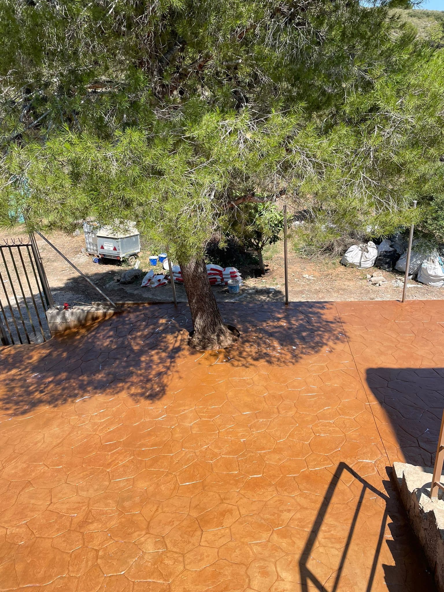 Patio de hormigón impreso en piedra rodena de tono anaranjado alrededor de un olivo, con la obra aún en curso al fondo.
