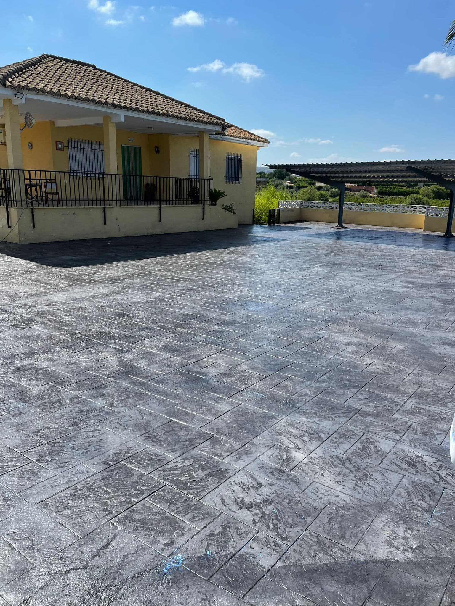 Gran explanada de hormigón impreso en piedra sillería gris ante una vivienda con porche y pérgola.
