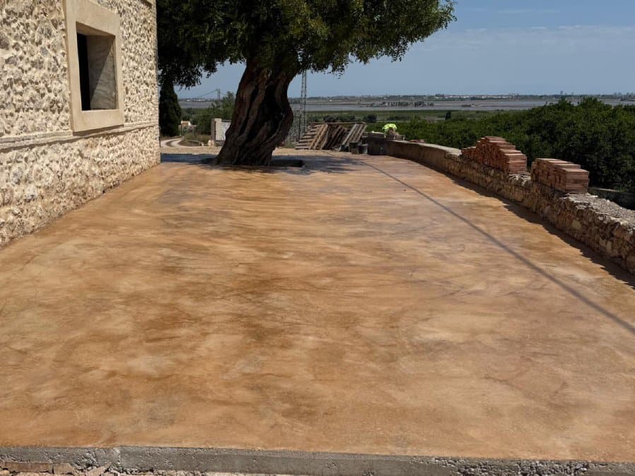Patio de hormigón fratasado de tono arena alrededor de un olivo, con el paisaje al fondo.