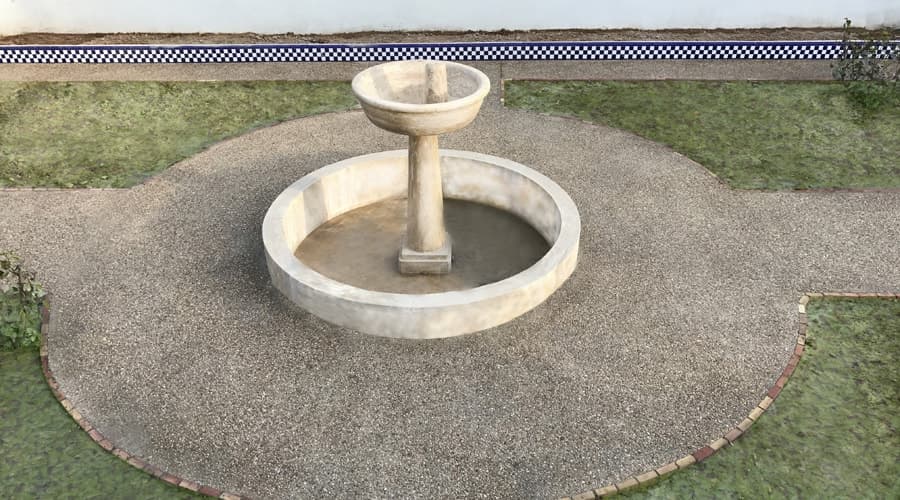Fuente circular rodeada de hormigón lavado con árido visto, con un zócalo de azulejo al fondo.