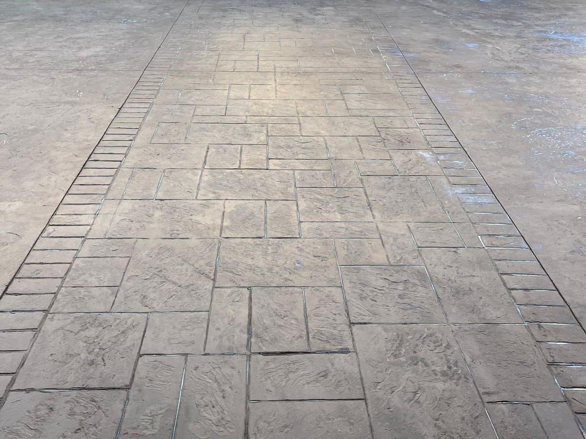 Pavimento de hormigón impreso gris con despiece rectangular y cenefa de adoquín en el borde.