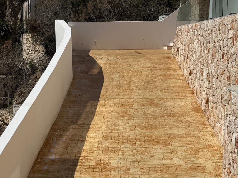 Rampa de acceso de hormigón impreso en adoquín pequeño de tono arena, entre un muro encalado y otro de piedra.