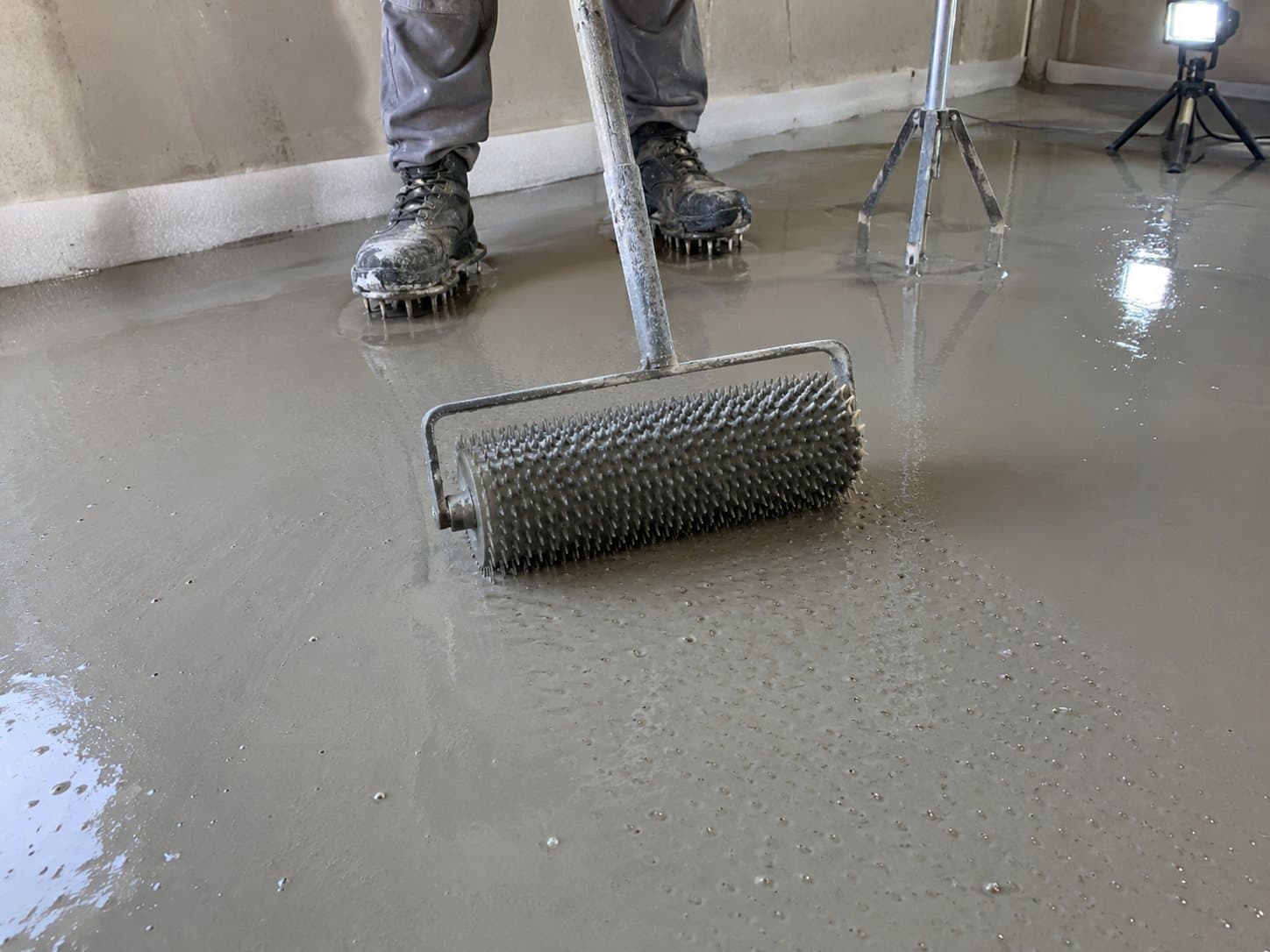 Rodillo de púas pasando sobre el autonivelante fresco, con las botas de púas del operario al fondo.