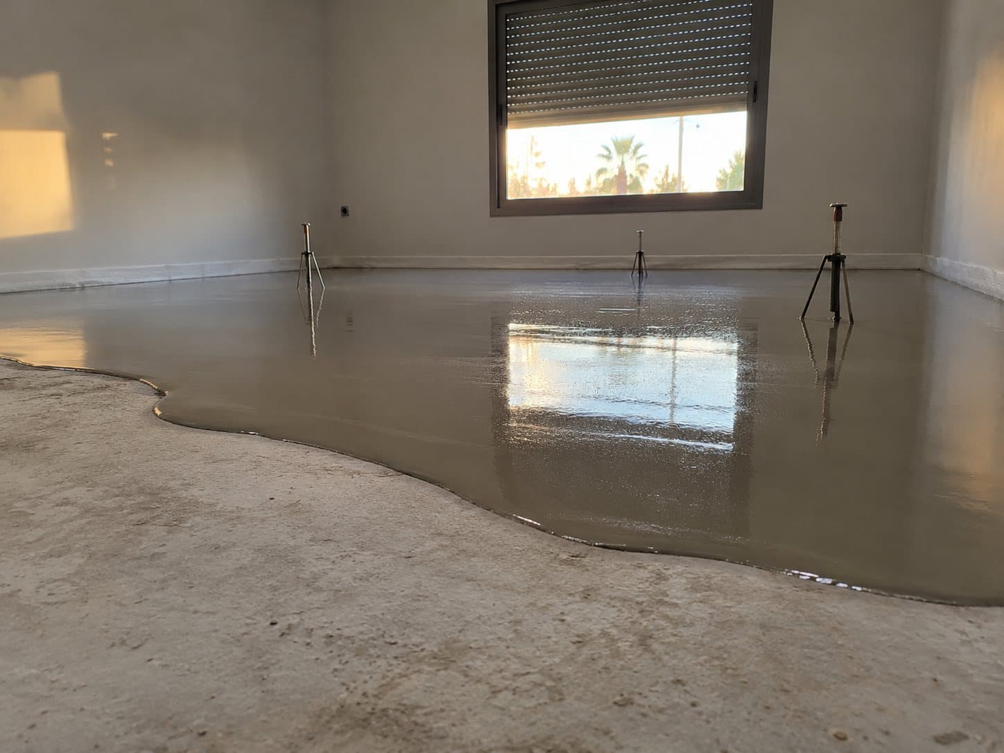Autonivelante recién vertido extendiéndose sobre el suelo de una habitación vacía, con trípodes de nivelación y la luz de la ventana reflejada.