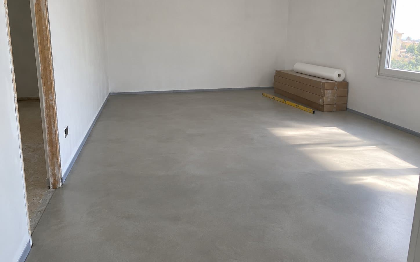 Habitación vacía con el suelo de autonivelante gris ya seco, cajas de tarima y un rollo de base aislante junto a la ventana.