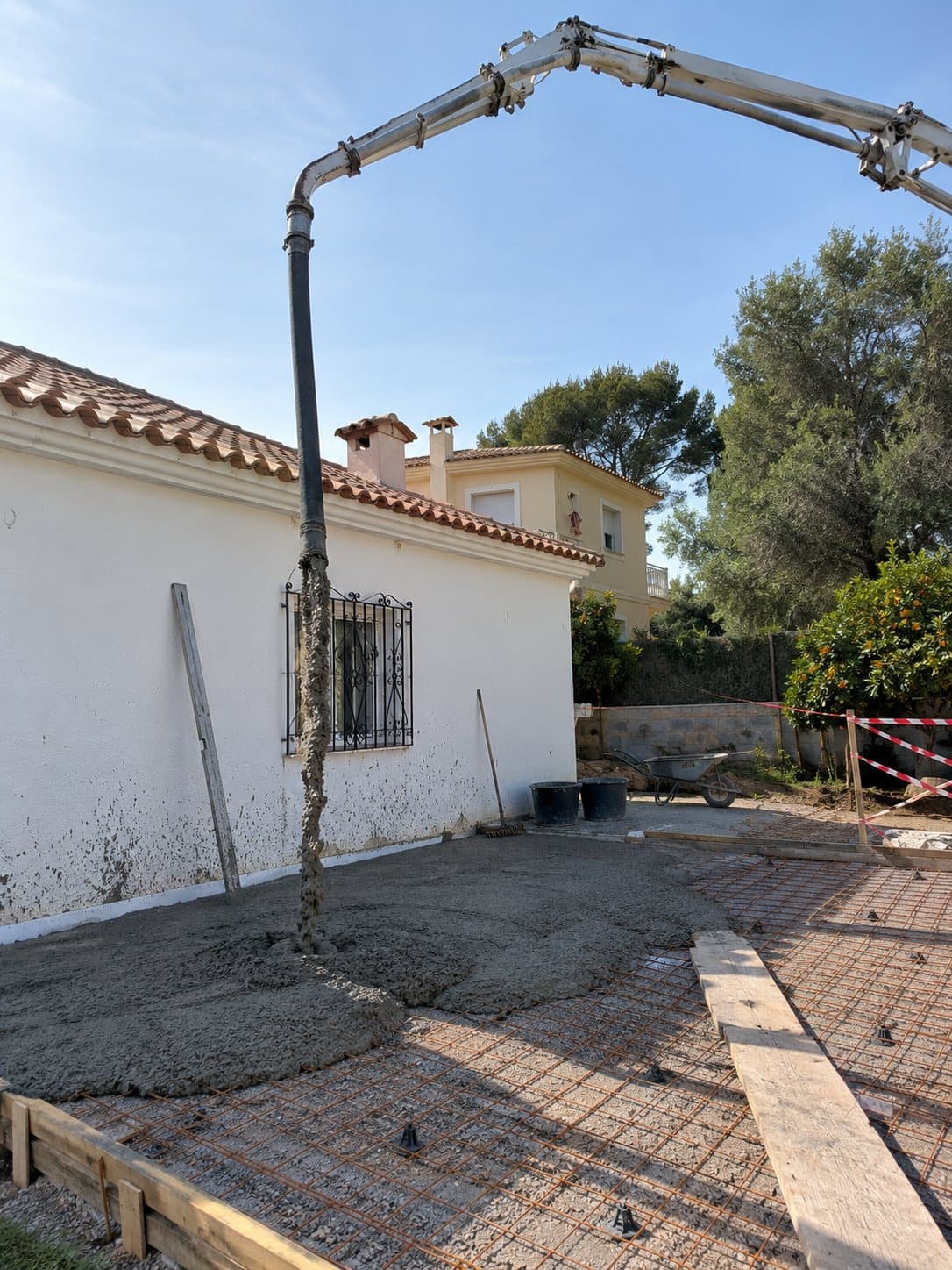 Pluma de bombeo vertiendo hormigón en el patio de una casa blanca de una planta, sobre el mallazo, entre encofrados de madera.