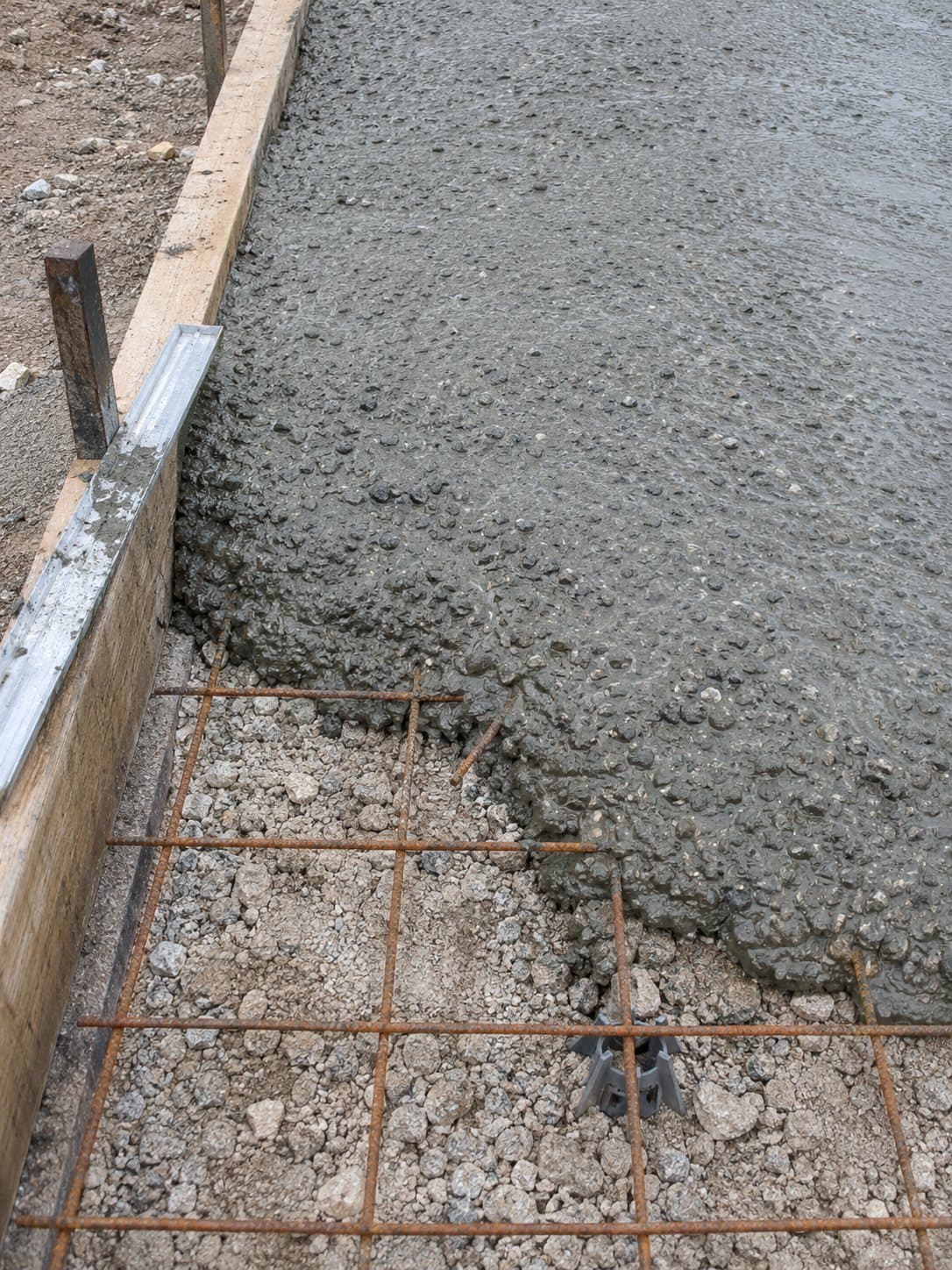 Hormigón recién vertido avanzando sobre el mallazo y la grava, junto a un encofrado de madera con regla de aluminio.