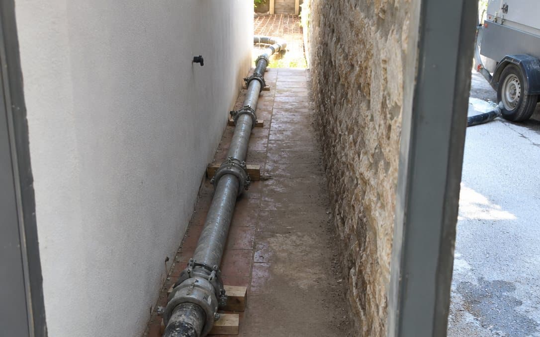 Tubería de bombeo de hormigón tendida por un pasillo estrecho entre la casa y un muro de piedra, hacia el patio trasero.