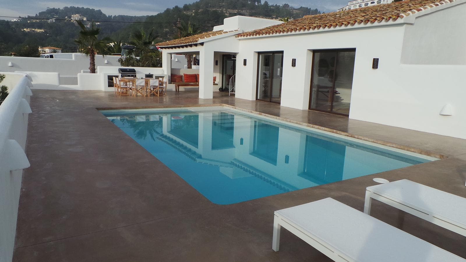 Contorno de piscina de hormigón continuo en tono tostado ante una vivienda encalada.