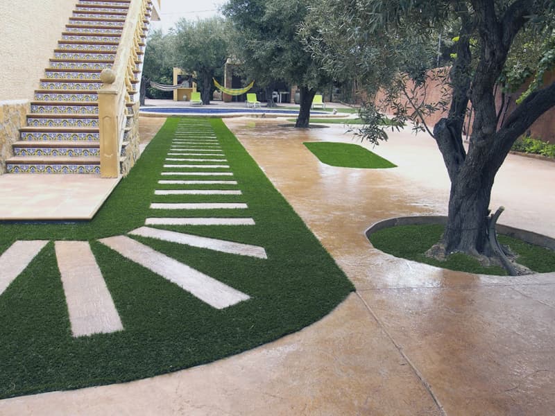 Jardín con olivos, césped y un pavimento de hormigón de tono ocre que rodea los alcorques.