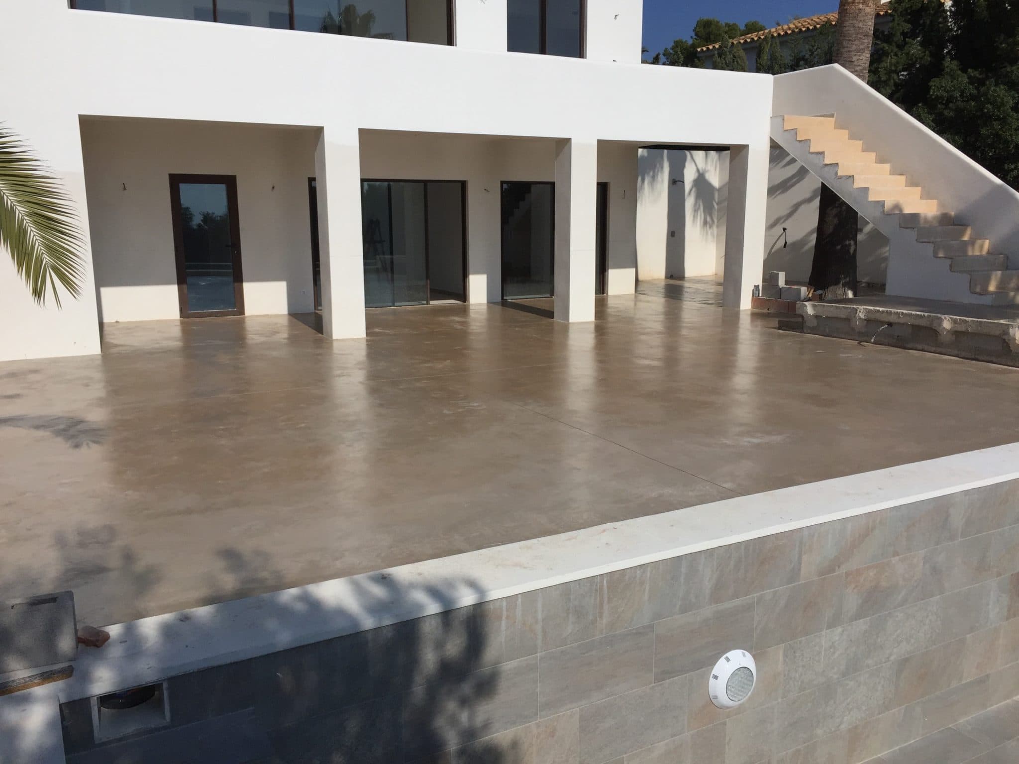 Terraza de hormigón pulido claro ante una vivienda blanca de líneas rectas.