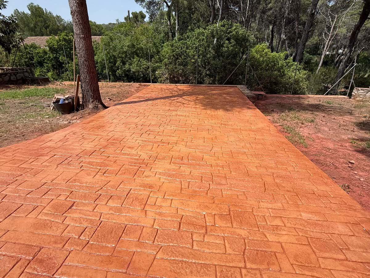Explanada de hormigón impreso en adoquín irregular de tono rojizo, entre pinos, con el terreno natural al fondo.