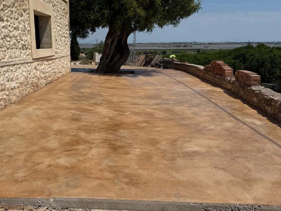 Patio de hormigón fratasado de tono arena alrededor de un olivo, con el paisaje al fondo.